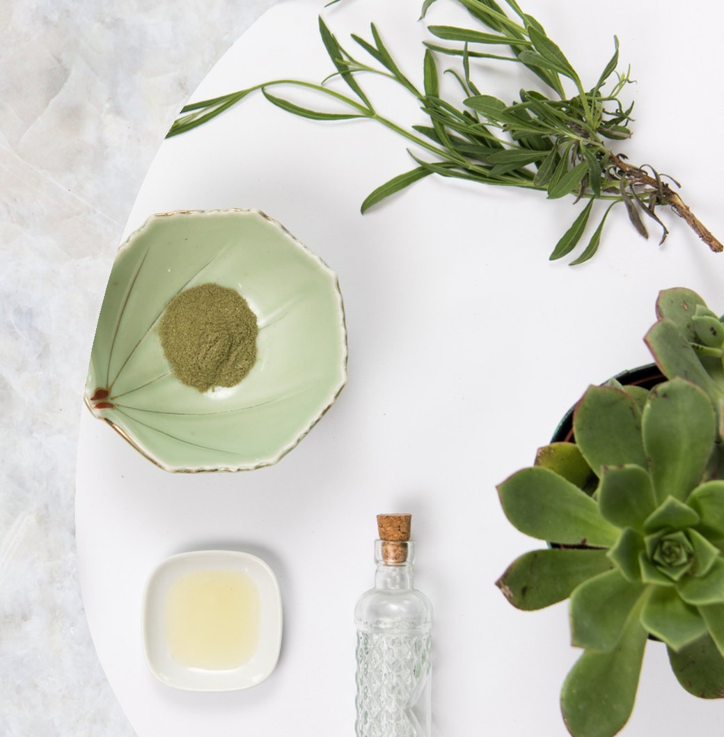 A celadon bowl of green botanical powder, pressed plant oil, a patterned glass vial and fresh herbs arranged on white marble — natural, organic Enigma Essentials skincare ingredients.